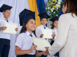Niña de primaria recibiendo diploma durante ceremonia escolar en Raíces del Saber.